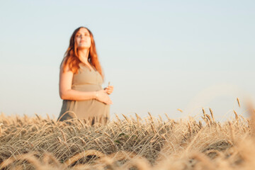 Fototapeta na wymiar beautiful young pregnant woman walks in ripe wheat field at sunset, expectant mother with red hair relax in nature stroking her belly with her hand