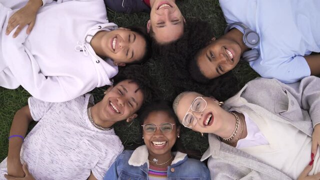 Relaxed teenager multiracial friends lying together on green grass with eyes closed, wake up and smile looking to camera