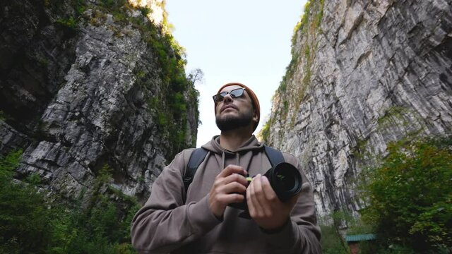 Young bearded tourist in sun glasses with camera taking pictures of wild mountain rocky nature. Adult traveler photographer on vacation trip in highland forest among rocks. High land natural tourism.