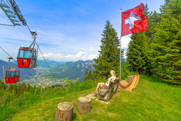 Red cable car cabin from Chur to Kanzeli and Brambruesch in Switzerland. Woman relaxing on top of Brambruesch. Swiss cable car of Chur or Coira with Swiss flag in Grisons canton.