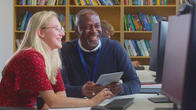 Female university or college student working at computer in library being helped by tutor holding digital tablet - shot in slow motion