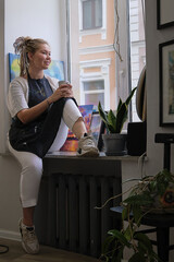 Artist Woman in dirty apron resting with a cup of tea sitting on the windowsill and looks out the window