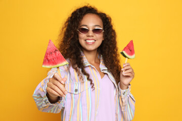 Beautiful young African American woman with pieces of watermelon against yellow background, focus on hand