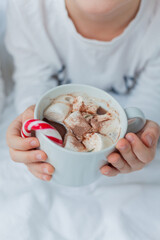 Adorable 6 year old boy in festive pajamas sits by the Christmas tree holding a mug of hot cocoa with marshmallows and lollipop in Christmas decorations. Winter holidays. New Year. Cozy Home.