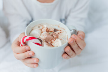 Adorable 6 year old boy in festive pajamas sits by the Christmas tree holding a mug of hot cocoa with marshmallows and lollipop in Christmas decorations. Winter holidays. New Year. Cozy Home.