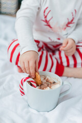 Adorable 2 year old girl in festive pajamas sits by the Christmas tree holding a mug of hot cocoa with marshmallows and lollipop in Christmas decorations. Winter holidays. New Year. Cozy Home.