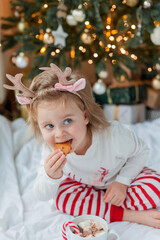 Adorable 2 year old girl in festive pajamas sits by the Christmas tree holding a mug of hot cocoa with marshmallows and lollipop in Christmas decorations. Winter holidays. New Year. Cozy Home.