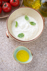 Bowl with italian stracciatella di bufala, green basil, olive oil and red tomatoes over beige marble background, elevated view, vertical shot
