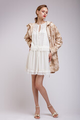 High fashion photo of a beautiful elegant young woman in a pretty jacket with beige streaks, white dress posing over soft gray background. Slim figure. Studio shot