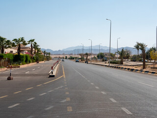 Fototapeta premium Safaga, Egypt - September 27, 2021: View of the main road stretching into the distance against the backdrop of mountains and blue sky. Cars drive along the highway to a neighboring city.