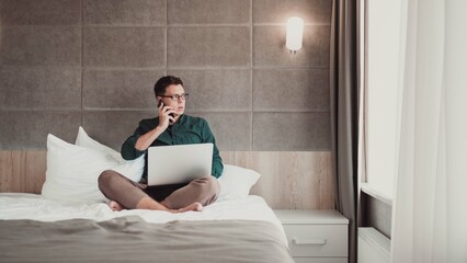 Young man working at home on laptop and talking on the phone,  looking out the window