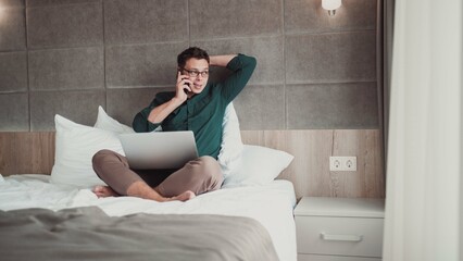 Young man working at home on laptop and talking on the phone,  looking out the window