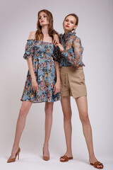 Two high fashion models in blue transparent blouse with floral pattern, dress, beige shorts. Beautiful young women. Studio shot. White background.  Slim figure. Make up, hairstyle. 