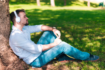 Young man wearing white headphones is listening to music on his smartphone and leaning on a tree in the city park.