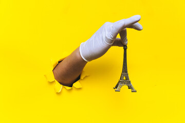 Fototapeta na wymiar A hand in a white medical glove holds a miniature metal Eiffel Tower. Torn hole in yellow paper. The concept of hygiene, self-isolation during quarantine the covid-19 coronavirus.