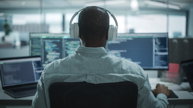 Office: Professional Black IT Programmer Uses Headphones while Working on Desktop Computer. Male Website Developer, Software Engineer Developing App, Video Game. Listening to Podcast, Music. Back View