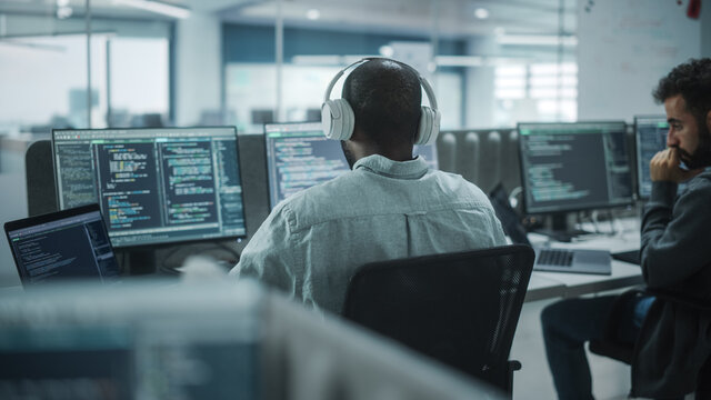 Office: Professional Black IT Programmer Uses Headphones while Working on Desktop Computer. Male Website Developer, Software Engineer Developing App, Video Game. Listening to Podcast, Music.
