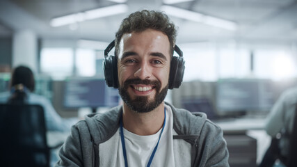 Office: Portrait of Happy IT Programmer Wearing Headphones Working on Desktop Computer, Looking at Camera and Smiling. Male Software Engineer Developing App, Video Game. Listening to Podcast, Music