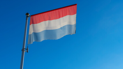 National flag of Luxembourg on a flagpole in front of blue sky with sun rays and lens flare. Diplomacy concept.