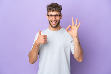 Fototapeta premium Delivery caucasian man isolated on purple background showing ok sign and thumb up gesture