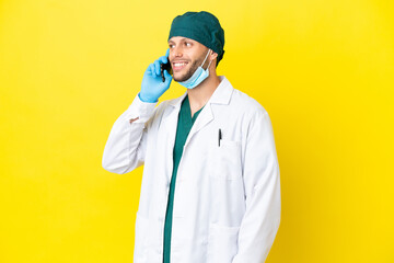 Surgeon blonde man in green uniform isolated on yellow background keeping a conversation with the mobile phone