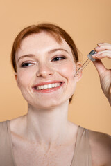Smiling red haired woman with freckles holding pipette with cosmetic serum isolated on beige