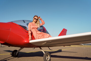 Pretty blond woman with wavy hair wearing pink silk classy dress posing near a private plane