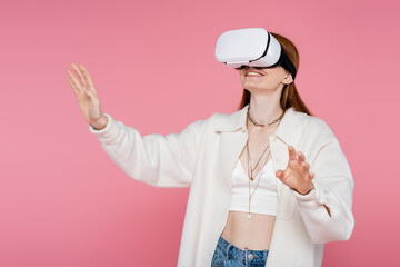 Cheerful and trendy woman in jacket gaming in vr headset isolated on pink