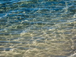 Texture of crystal clear light blue and turquoise transparent water with fine white sand base with bright reflection of sun in water movement. Water background