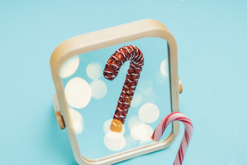 A red and white candy cane in a mirror reflecting a festive Christmas bauble ornament version of himself. Minimal creative New Year holiday layout with copy space on blue background. Self identity.