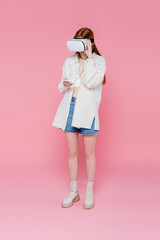 Full length of red haired woman in vr headset using smartphone on pink background