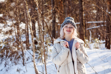 Fototapeta na wymiar Smiling blonde woman in warm knitted cap and beige coat outdoors in winter forest