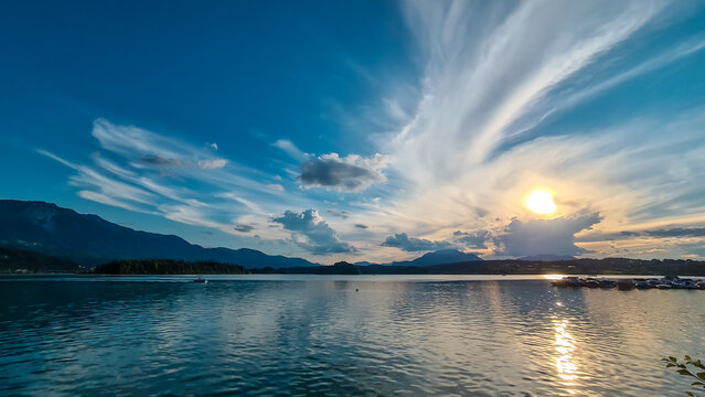 A panoramic view on the Lake Faak in Austria. The lake is surrounded by high Alpine peaks. The sun in slowly setting behind the mountains. Lots of clouds. Calm surface reflects the sunbeams. Serenity
