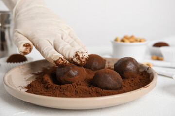Confectioner covering delicious chocolate truffles with cocoa powder at white table, closeup