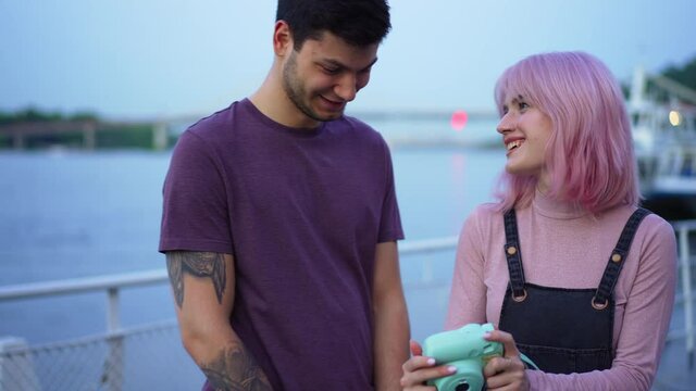 Smiling loving boyfriend admiring photos in camera as cheerful talented girlfriend showing pictures. Front view millennial Caucasian couple dating on urban embankment outdoors talking laughing
