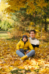 Happy young mother and little son sitting in autumn park among the yellow maple leaves