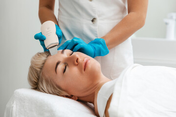 Naklejka premium Professional care. A cosmetologist in blue gloves does an ultrasound cleaning of a woman's face. Cleansing and rejuvenating procedure in a beauty salon. Side view