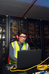 Obraz premium Asian male engineer wearing ear plugs using a laptop in computer server room