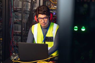 Asian male engineer wearing ear plugs using a laptop in computer server room