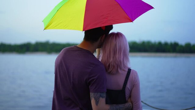 Back view young tattooed man hugging slim woman dating on embankment admiring beauty of river. Happy Caucasian boyfriend and girlfriend enjoying leisure outdoors with colorful umbrella