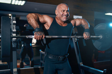 Fototapeta na wymiar Emotional adult bald bodybuilder doing trapezius and deltoid exercise with a barbell in the gym