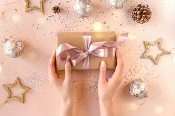 Female hands with natural manicure holding present in kraft paper with pink ribbon on soft beige background. Xmas composition. Flat lay. Happy holidays, New Year celebration and giving love concept. 