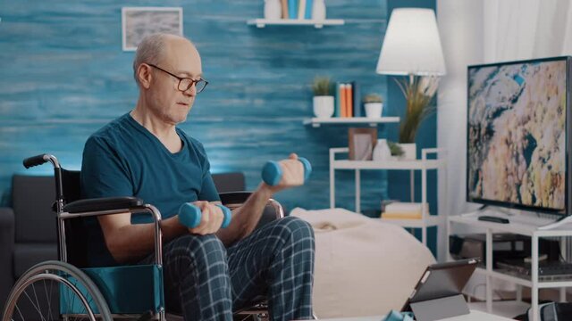 Aged man sitting in wheelchair and lifting weights while following video of workout exercise. Senior person with physical disability using dumbbells and watching training lesson on tablet