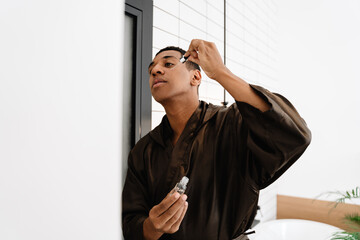 Black man wearing bathrobe applying face serum in bathroom