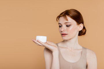 Young redhead woman looking at jar with cosmetic cream isolated on beige