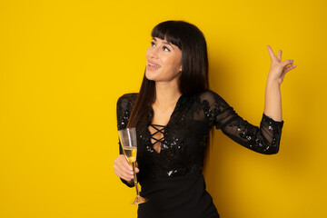 Young celebrating woman black dress . Beautiful model portrait isolated over yellow background hold wine glass.