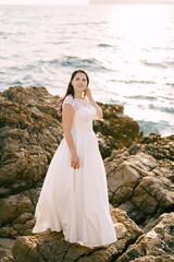 Fototapeta na wymiar Bride in a white lace dress stands on a rocky seashore by the sea