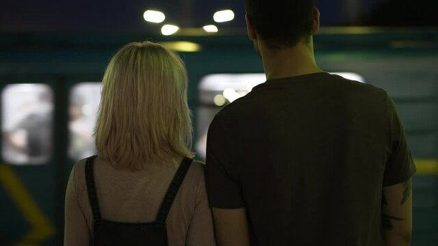 Back view Caucasian young couple standing on metropolitan station as train passing at background leaving. Man and woman dating outdoors in urban city at night waiting for transport. Slow motion
