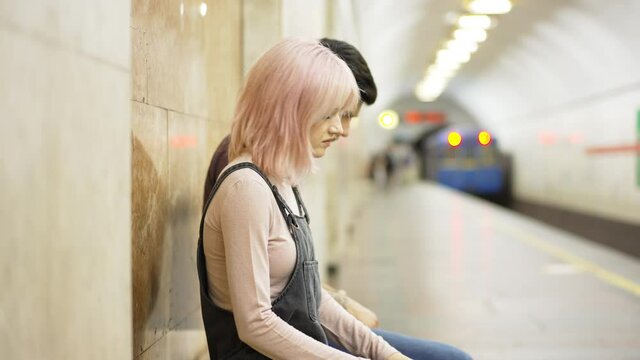 Side view young couple sitting indoors on metro station as train leaving in slow motion. Medium shot relaxed Caucasian millennial man and woman waiting for transport in urban city. Lifestyle