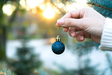 Small blue Christmas ball in a female hand against the background of Christmas trees